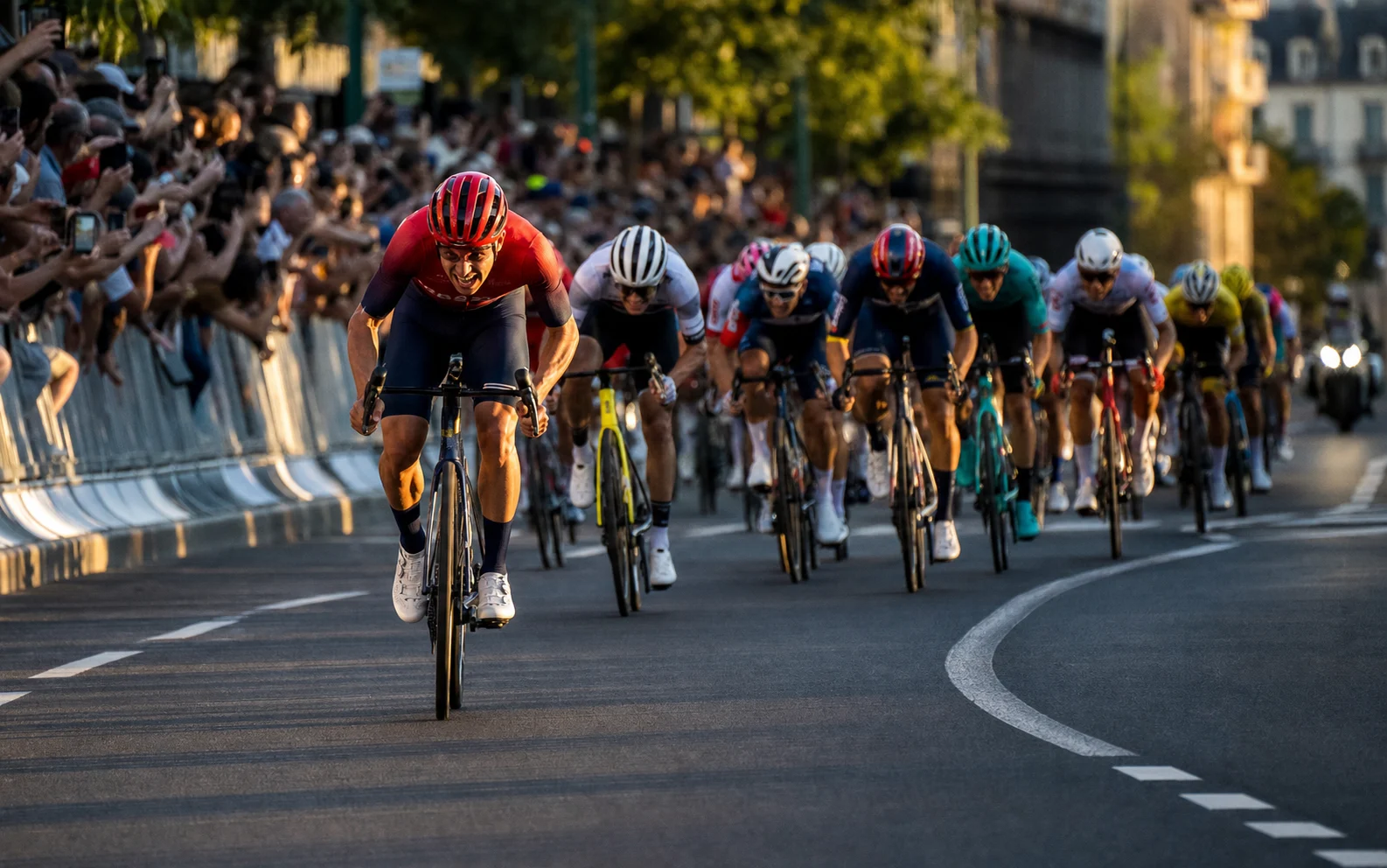 Un cycliste en tenue rouge lance un sprint prolongé devant un peloton compact dans une avenue française